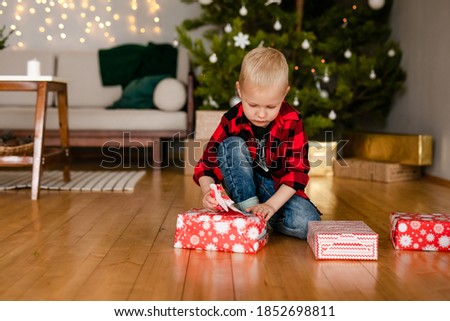 Little blonde boy cozy clothes and Santa Claus hats unpack gifts at home in the new year's interior.