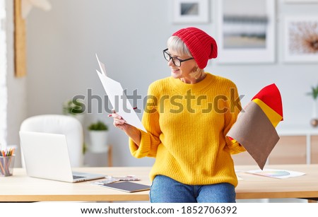 cheerful old woman creative designer in a red hat smiles at  workplace at the table with a laptop, sketches and samples
