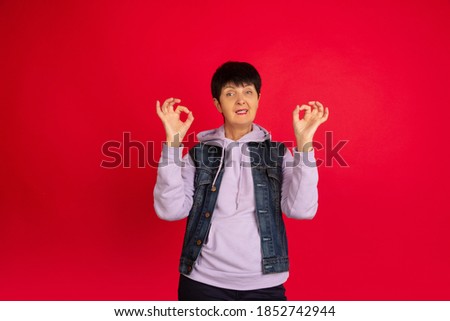 Nice sign Portrait of senior woman in stylish outfit, attire isolated on red studio background. Tech and joyful elderly lifestyle concept. Trendy colors, forever youth. Copyspace for your ad.