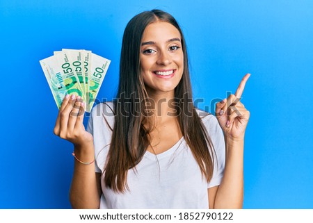 Young hispanic woman holding israel shekels smiling happy pointing with hand and finger to the side 