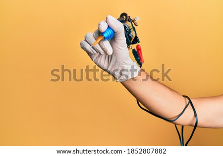 Hand of young hispanic man using tattoo machine over isolated yellow background.