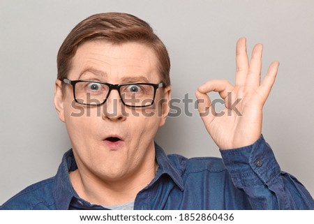 Studio close-up portrait of amazed impressed blond mature man with glasses, showing okay gesture with one hand, expressing his approval and acceptance. Headshot over gray background