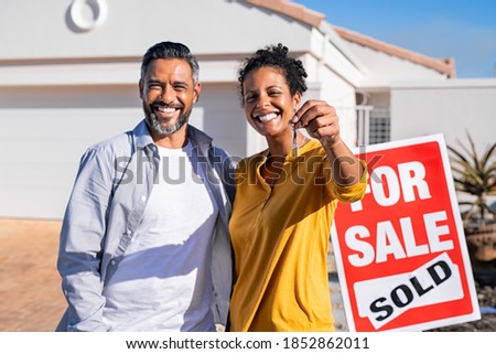 Portrait of happy mixed race couple holding house keys standing near sold signboard. Middle eastern man embracing african woman while showing house key outside of their new home. Moving home day.