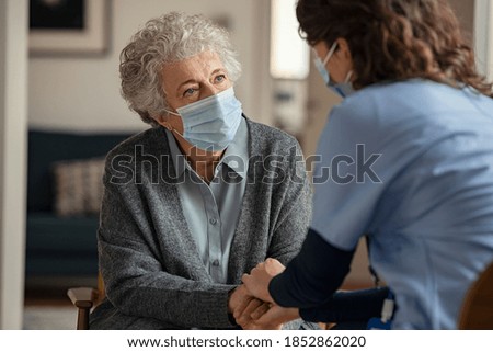 Elderly woman talking with a doctor while holding hands at home and wearing face protective mask. Worried senior woman talking to her general practitioner visiting her at home during virus epidemic.  Royalty-Free Stock Photo #1852862020