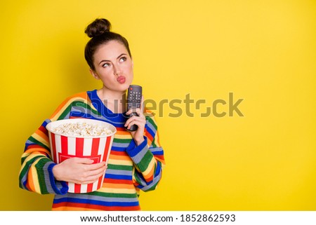 Photo of young attractive woman think hold remote control eat pop-corn look empty space isolated over yellow color background
