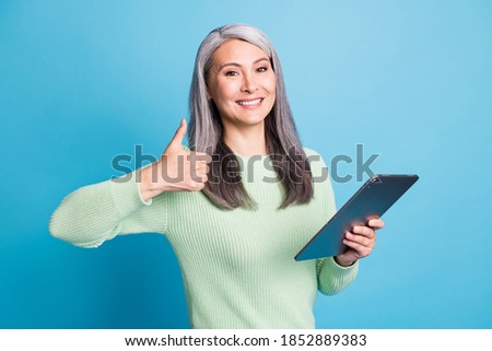 Photo of positive old lady hold tablet show thumb up sign wear green jumper isolated over blue pastel color background