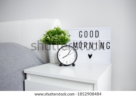 good morning concept - close up of alarm clock, houseplant and lightbox with "good morning" words on bedside table in bedroom