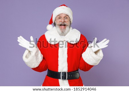 Shocked elderly Santa Claus man in Christmas hat red suit coat glasses keeping mouth open spreading hands isolated on violet purple background studio. Happy New Year celebration merry holiday concept