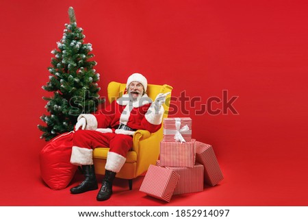 Surprised Santa Claus man in Christmas hat suit sit in armchair with fir tree presents gifts pointing index finger aside isolated on red background. Happy New Year celebration merry holiday concept