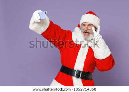 Blinking Santa Claus man in Christmas hat red suit coat glasses doing selfie shot on mobile phone showing victory sign isolated on violet background. Happy New Year celebration merry holiday concept