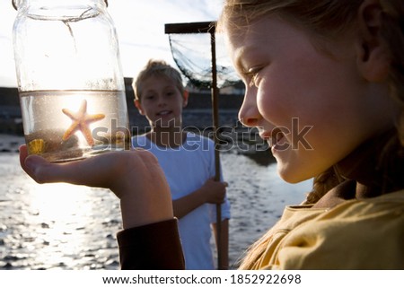 Close up of a little girl holding up a jar containing a starfish kept in water while her brother is watching her in the background