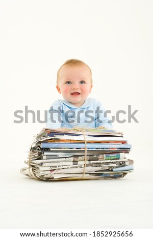 Vertical portrait of a baby boy sitting by a bundle of newspapers smiles at the camera.