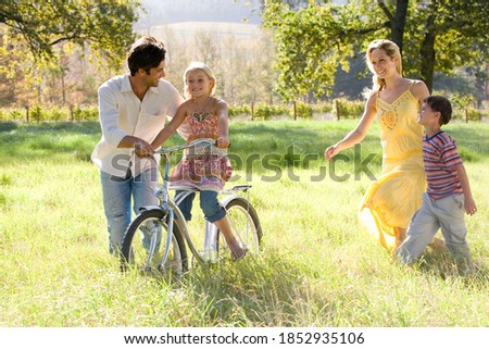 Horizontal shot of a cheerful family of four in field with father helping daughter on the bicycle as mother and son looks on.