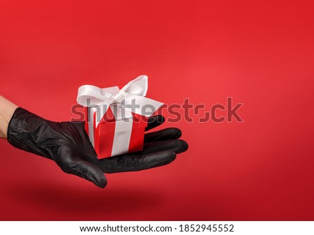 A gift box decorated with a bow lies in the palm of your hand in a black protective glove on a red background with copy space. Safe communication.