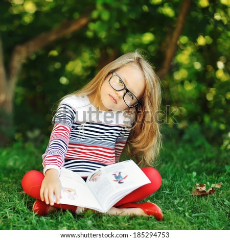 Adorable little girl with book in a summer park wearing glasses 