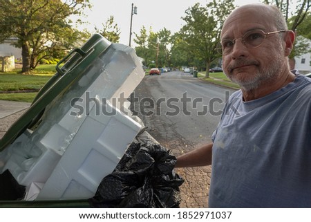 Bald senior citizen man standing in a street with his hand on a full trash can