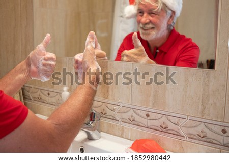 Coronavirus. A happy senior man in santa hat washes his hands at home gesturing ok sign with his hand. Red surgical mask to prevent coronavirus infection on the sink