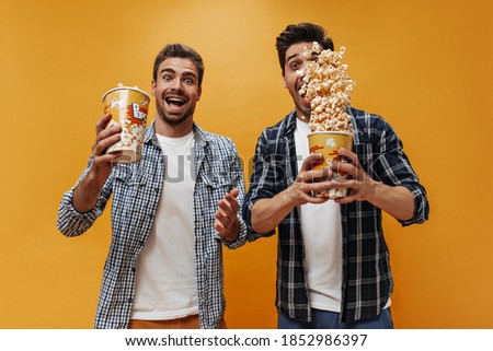 Emotional crazy bearded men in checkered shirts scatter popcorn on isolated. Cool guys in white t-shirts rejoice on orange background.