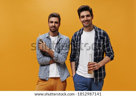 Portrait of handsome bearded brunet men in white t-shirts, checkered shirts and colorful shorts smile and hold beer bottles on orange background.