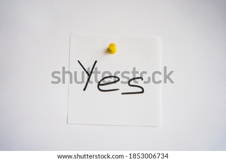 White note paper with word YES written in black handwriting pinned on white paper background with yellow pin. Business note.