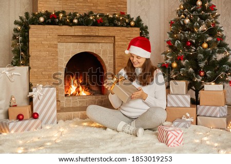 Lovely woman opening gift on Christmas, sitting on floor near fireplace and tree, smiling female wearing santa hat, white sweater and warm socks, sitting with crossed lags and holding present box.