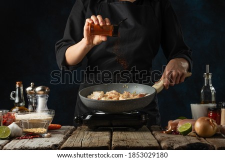 Professional chef pours hot spices to pan wok with frying chicken. Backstage of cooking traditional Indian chicken curry on dark blue background. Frozen motion. Concept of cooking tasty hot meal.
