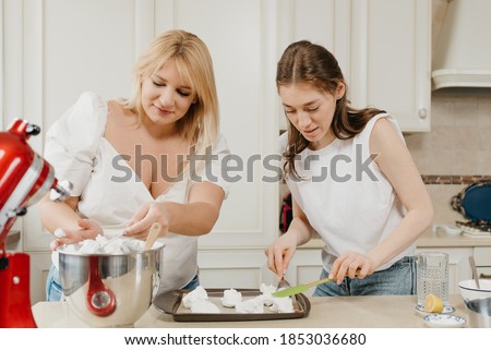 Two smiling young women are putting diligently the whipped meringue on a tray with a spoon and a scapula in the kitchen. Girls are preparing to cook a delicious lemon meringue tart.