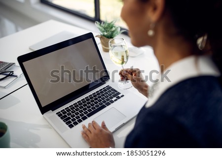 Close-up of businesswoman having a glass of wine while surfing the net on laptop in the office. Focus is on laptop's screen. Copy space. 