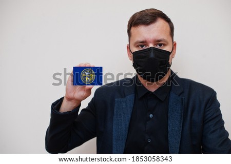 Man wear black formal and protect face mask, hold Nebraska flag card isolated on white background. USA coronavirus Covid country concept.