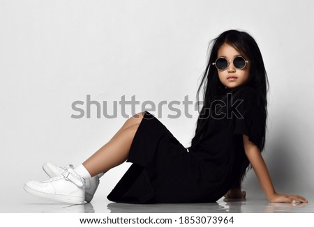 Full-growth portrait of cool asian kid girl in stylish black dress, round sunglasses and white sneakers sitting on floor side to camera leaning back on her hands. Trendy children fashion concept Royalty-Free Stock Photo #1853073964