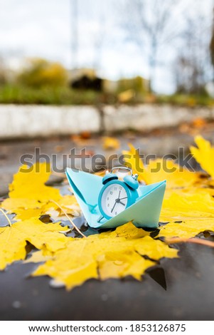 blue alarm clock floating on a paper ship over water surface with autumn leaves. wasting time concept. time to travel conceptual.