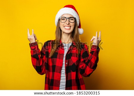 Young woman in santa hat and red plaid shirt makes rock and roll gesture on yellow background