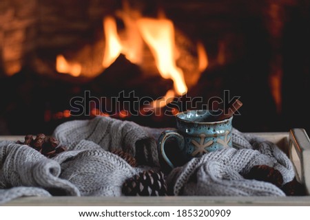 A cup of hot chocolate with cinnamon stick on a tray with warm scarf in front of a fireplace
