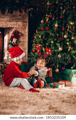 Brother and sister are sitting with boxes in green packaging with gifts and a bow. Christmas mood. Children in New Year's costumes waiting for a miracle. Against the background of a Christmas tree and