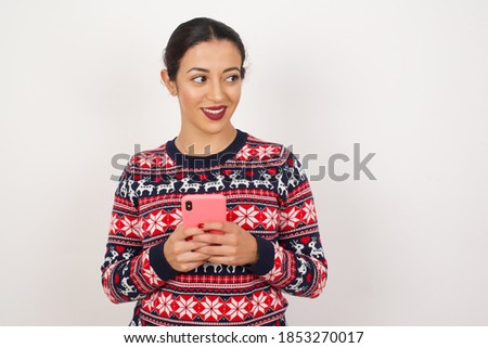 Beautiful Arab girl wearing Christmas sweater over isolated white background hold telephone hands read good youth news look empty space advert 