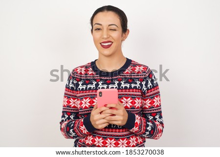 Portrait of Beautiful Arab girl wearing Christmas sweater over isolated white background, taking a selfie  celebrating success