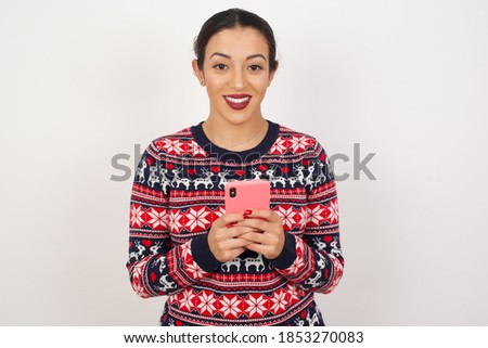 Photo of Beautiful Arab girl wearing Christmas sweater over isolated white background enjoys distant communication, uses mobile phone, surfs fast unlimited internet, has pleasant smile, makes shopping