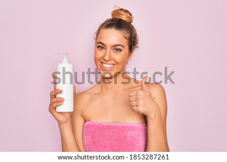 Beautiful woman with blue eyes wearing towel shower after bath holding bottle of body cream happy with big smile doing ok sign, thumb up with fingers, excellent sign