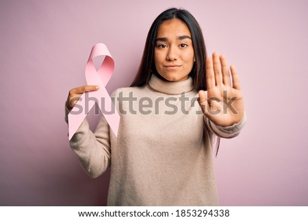 Young beautiful asian woman holding pink cancer ribbon symbol over isolated background with open hand doing stop sign with serious and confident expression, defense gesture