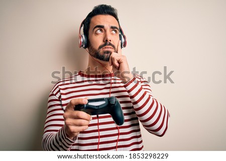 Young handsome gamer man with beard playing video game using joystick and headphones serious face thinking about question, very confused idea