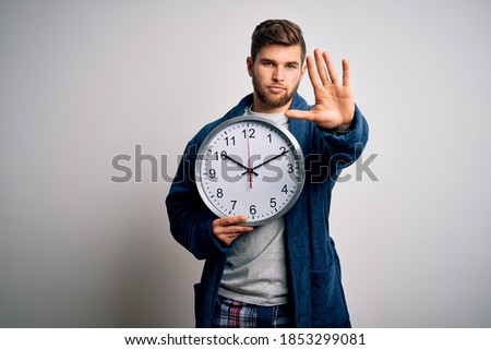 Young blond man with beard and blue eyes wearing pajama holding big clock with open hand doing stop sign with serious and confident expression, defense gesture