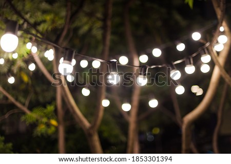 Blurred background, backyard illumination, light in the evening garden, electric lanterns with round diffuser. Lamp garland of light bulbs on a tree branch among the leaves, illuminate night scene. Royalty-Free Stock Photo #1853301394
