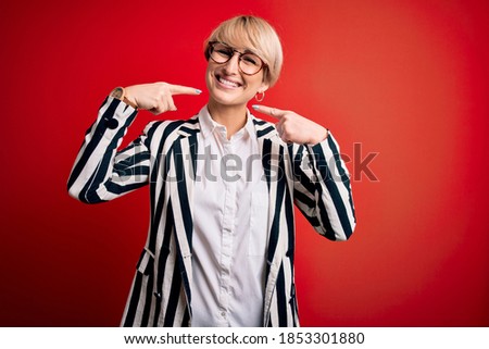 Blonde business woman with short hair wearing glasses and striped jacket over red background smiling cheerful showing and pointing with fingers teeth and mouth. Dental health concept.