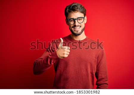 Young handsome man with beard wearing glasses and sweater standing over red background doing happy thumbs up gesture with hand. Approving expression looking at the camera showing success.