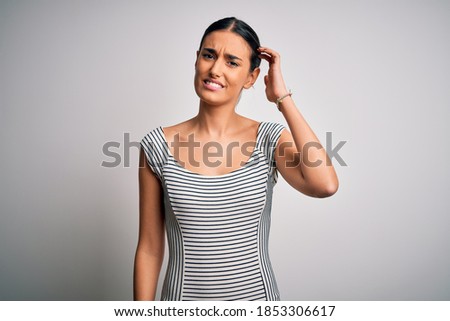 Young beautiful brunette woman wearing casual striped dress over isolated white background confuse and wonder about question. Uncertain with doubt, thinking with hand on head. Pensive concept.