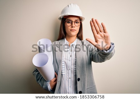 Young beautiful architect woman wearing safety helmet and glasses holding blueprints with open hand doing stop sign with serious and confident expression, defense gesture
