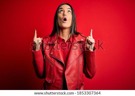 Young beautiful brunette woman wearing casual jacket standing over isolated red background amazed and surprised looking up and pointing with fingers and raised arms.