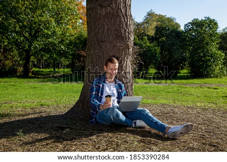 The guy sits near a tree in the park with a cup of coffee and works on a laptop. Freelance work outdoors. Working outside the office due to the Covid 19 pandemic
