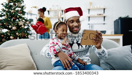 Close up of happy African American little kid sitting with dad in santa hat at decorated room and watching cartoons on tablet. Mom and child decorating Christmas tree on background. Xmas concept