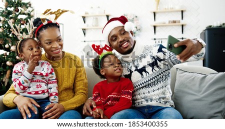Portrait of happy African American family taking pictures on smartphone and smiling in cozy christmassy decorated home. Dad in santa hat taking selfie photo on cellphone with kids and wife